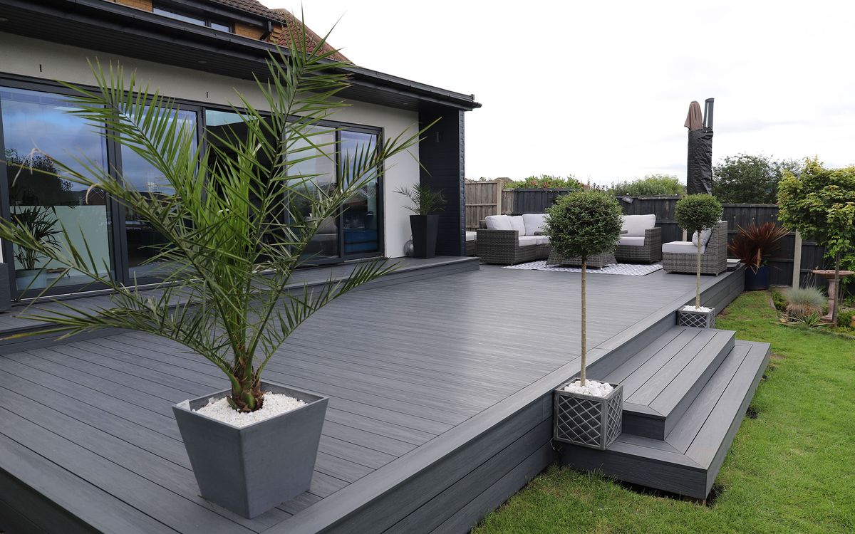 Modern outdoor terrace with warm grey composite decking, woven lounge chairs, black glass doors, railing and forest greenery.